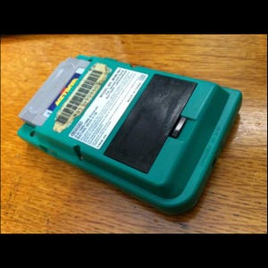 Könnte beinhalten: Ein teal Nintendo Game Boy mit eingelegter Metroid-Spielkassette. Der Game Boy hat eine schwarze rechteckige Abdeckung und einen silbernen Schalter. Die Spielkassette hat ein blaues und gelbes Etikett. Auf einer Holzoberfläche.