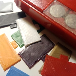 May include: A collection of colorful, rectangular plastic pieces with a textured surface and a small tab. The pieces are in various colors, including white, orange, purple, and green. A red plastic object is in the background.