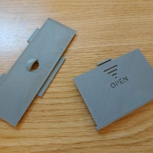 May include: Two grey plastic rectangular pieces. One has a circular opening and two side tabs. The other has the word "OPEN" printed on it, along with a graphic of three lines and a tab.