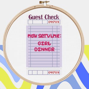 May include: Cross-stitch embroidery in a wooden hoop. The design features a mock guest check with the text "Guest Check" and "now serving: GIRL DINNER" in pink. The background has a colorful, abstract design.