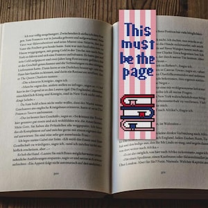 May include: Open book with a bookmark. The bookmark has a pink and white striped background with the text "This must be the page" in blue. Below the text are three stacked books.