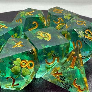 May include: Set of seven polyhedral dice with a green, transparent appearance. Each die features gold-colored numbers and clover designs. The dice are designed for tabletop role-playing games.