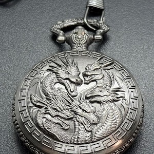 May include: Silver pocket watch with a detailed design of four dragons on the face. The watch has a decorative chain attached.