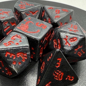 May include: A set of seven polyhedral dice in a dark, metallic charcoal gray finish with bright red engraved numbers and dinosaur-themed icons. The collection includes various shapes such as a d20, d12, d10, d8, d6, and d4, all featuring sharp edges.