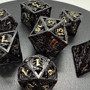 May include: Set of seven black metal dice with gold numbering and intricate filigree designs. The dice are in various geometric shapes, including a d20, d12, d10, d8, d6, and d4, suitable for tabletop role-playing games.