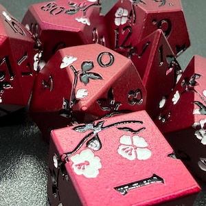 May include: Close-up of a collection of red polyhedral dice. Each die features black numbers and is decorated with a floral design in black and white. The dice are arranged in a pile, showcasing their intricate details.