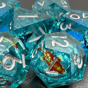 May include: A set of polyhedral dice with a clear, turquoise resin. Each die features silver numbering and contains miniature koi fish and aquatic elements. The dice are designed for tabletop gaming.