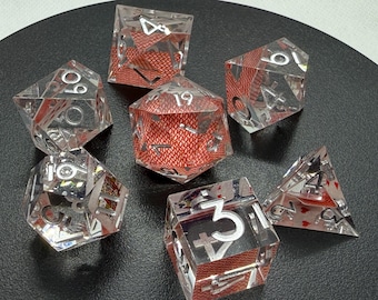 Puede incluir: Juego de siete dados de resina transparente con numeración plateada y diseños de naipes rojos en su interior. Los dados tienen varias formas geométricas, incluyendo un d4, d6, d8, d10, d12 y d20. Los dados están sobre una superficie negra.