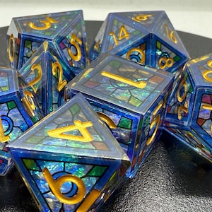 Puede incluir: Juego de siete dados poliédricos con diseño de vidriera. Los dados son azules con números dorados y un patrón de mosaico colorido. Ideales para juegos de mesa, estos dados son una adición única a cualquier colección.
