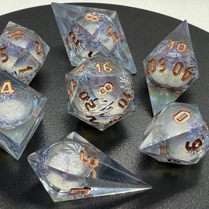 Puede incluir: Juego de siete dados poliédricos con resina transparente e iridiscente. Cada dado presenta un núcleo esférico con un diseño blanco brillante y números de color cobre. Los dados están dispuestos sobre una superficie oscura.