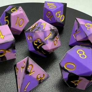 May include: A collection of seven multi-sided dice in purple, black, and pink hues. Each die has gold numerals. These dice are designed for role-playing games and are presented on a dark surface.