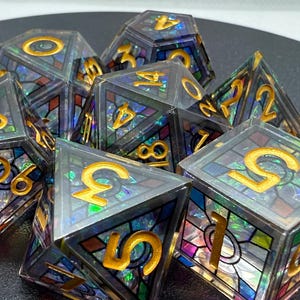 May include: A set of polyhedral dice with a stained glass design. The dice are clear with gold-colored numbers and colorful, iridescent panels. The dice are arranged in a pile, showcasing their various shapes and facets.