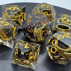 Puede incluir: Un conjunto de siete dados transparentes con purpurina dorada y anillos dorados incrustados. Los dados están numerados del 1 al 6 y están dispuestos sobre una superficie negra.