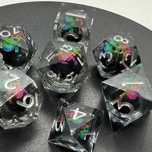 May include: Set of seven clear iridescent dice with silver numbers and a black and rainbow iridescent design. The dice are on a black surface.