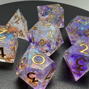 May include: Set of seven iridescent dice with gold numbers. The dice are made of clear resin with purple and gold flecks. The dice are in various shapes, including a d4, d6, d8, d10, d%, d12, and d20.