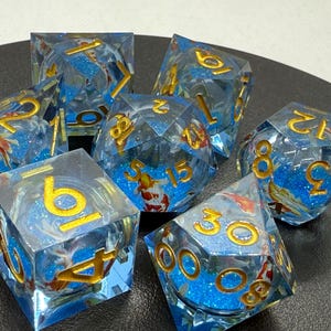 May include: A set of seven blue and gold dice with a glittery finish. The dice have a variety of numbers and symbols on them, including a koi fish design.