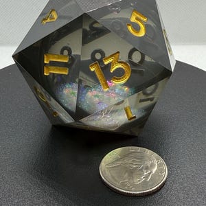 May include: A large, clear, multi-sided die with gold-colored numbers, filled with iridescent glitter. A U.S. quarter coin is next to the die for size comparison. The die is set against a dark background.