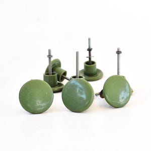 Pode incluir: Um conjunto de botões verde-oliva com um acabamento liso e brilhante. Cada botão apresenta um topo circular e uma haste de metal que se estende a partir da parte de trás, concebida para fixação ao tecido. O conjunto inclui várias peças.
