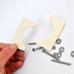 May include: Two off-white plastic pipe clamps with a rounded opening. The clamps are separated and surrounded by silver screws and washers.