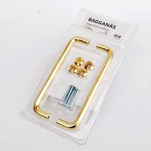 Może przedstawiać: Opakowanie złotych uchwytów do szafek, okuć i śrub. Uchwyty są prostokątne z zaokrąglonymi narożnikami. Opakowanie jest oznaczone napisem "BAGGANÄS". Uchwyty mają około 12.7 cm długości.