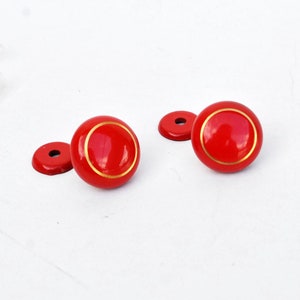 May include: Two red round cabinet knobs with a gold trim. The knobs are on a white surface.