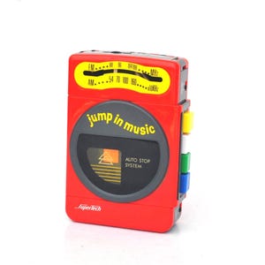 Puede incluir: Un reproductor de casetes portátil rojo con el texto "jump in music" y un dial de radio AM/FM. El reproductor tiene un área circular gris oscuro con una ventana de casete y varios botones de colores en el lateral.