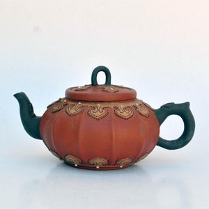 Puede incluir: Una tetera de arcilla roja con cuerpo redondo y asa negra. La tetera está decorada con detalles dorados y diseños intrincados.