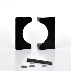May include: Two black, square-shaped brackets with a circular opening, positioned side-by-side. A metal rod, washers, and nuts are below the brackets. The items are on a white surface.
