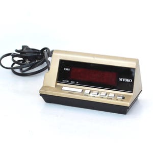 Puede incluir: Un reloj despertador digital vintage con una parte superior dorada y una base negra. El reloj muestra dígitos rojos y tiene varios botones debajo de la pantalla. La marca "MYOKO" es visible. Un cable de alimentación negro está conectado.
