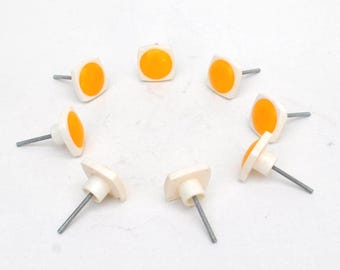 May include: A set of eight square cabinet knobs arranged in a circle. Each knob features a white square base with a rounded orange circular center and a silver metal screw extending from the back for mounting purposes on furniture or cabinetry.