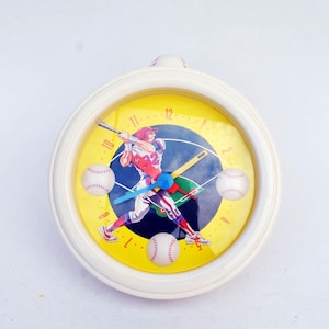 May include: A round, white baseball-themed clock with a yellow face. The clock face features a baseball player swinging a bat, baseballs, and blue, green, and red clock hands. The clock has a white frame and is designed to be hung on a wall.