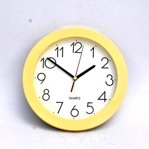 May include: A small, round quartz clock with a pale yellow frame. The clock face is white with black numerals and hands. The word "QUARTZ" is printed below the center. The clock is approximately 10 cm in diameter.