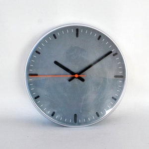 May include: A round, grey clock with black hour and minute hands and an orange second hand. The clock has a silver frame.