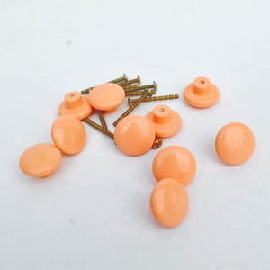 Może przedstawiać: Kolekcja brzoskwiniowych gałek do szafek i małych mosiężnych śrubek rozrzuconych na białej powierzchni. Gałki mają zaokrąglony, owalny kształt i błyszczące wykończenie. Śruby są cienkie i mają złoty odcień.