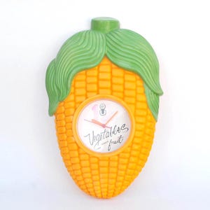 May include: A novelty wall clock shaped like an ear of corn. The clock has a yellow body with green leaves and a white clock face. The clock face has red hands and the words "Vegetables & fruit".