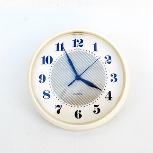May include: A white analog clock with black numbers and blue hands. The clock face has a silver, textured background and the words "QUARTZ" printed on it.
