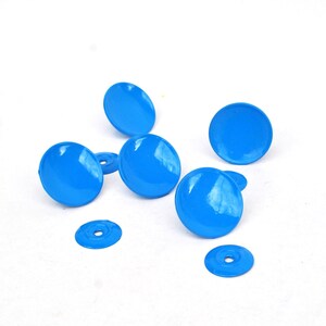 Peut inclure: Des boutons de meuble ronds bleu vif et des rondelles sont dispersés sur une surface blanche. Les boutons ont une finition brillante et sont espacés uniformément. Les rondelles sont plates avec un trou central. Quincaillerie.