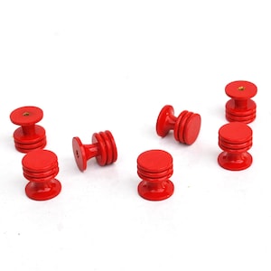 Puede incluir: Colección de siete pequeños pomos de madera rojos. Cada pomo tiene un cuerpo cilíndrico con una parte superior e inferior circulares. Los pomos están espaciados sobre un fondo blanco, mostrando su diseño.