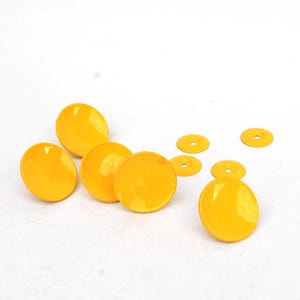 May include: A collection of bright yellow buttons and washers are scattered on a white surface. The buttons are oval-shaped and glossy, while the washers are small and circular with a hole in the center. These could be used for sewing or crafting.