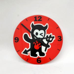 May include: Red wall clock with a black cartoon devil holding a pitchfork. The clock face has black numbers 12, 9, 3, and 6.