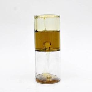 Könnte beinhalten: Ein Spielzeug mit flüssiger Bewegung aus klarem Glas in Form einer Sanduhr. Der obere und mittlere Abschnitt sind mit einer gelbbraunen Flüssigkeit gefüllt, während der untere Abschnitt leer ist. Die Flüssigkeit fließt langsam von oben nach unten.