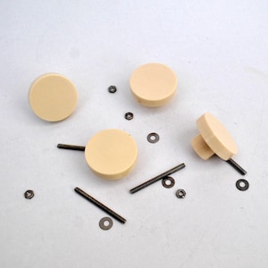Puede incluir: Cuatro pomos redondos de color beige para armario con tornillos y arandelas de metal. Los pomos están sobre una superficie blanca.