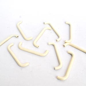 May include: Several off-white, curved metal handles, each with a small hole at the end, are scattered on a white surface. These handles are likely for furniture or cabinets.