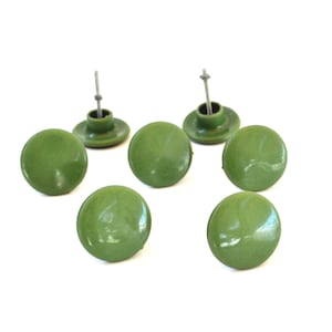 Puede incluir: Un conjunto de seis botones redondos de color verde oliva con un acabado liso y brillante. Cada botón presenta una parte superior plana y circular, con un vástago de metal en la parte inferior para su fijación. Los botones están agrupados.