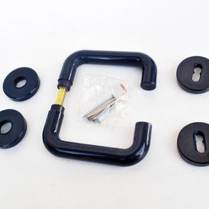 May include: A set of black door hardware, including a pair of curved handles, four round escutcheons, and a packet of screws. The handles have a brass-colored connection point. The hardware is set against a white background.