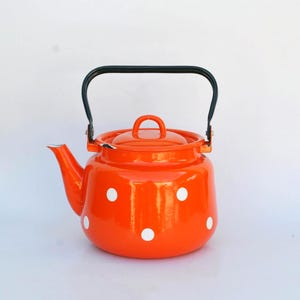 May include: Orange enamel teapot with white polka dots. The teapot has a black handle and a lid with a small handle. The spout is curved, and the teapot is set against a white background. A classic kitchenware item.