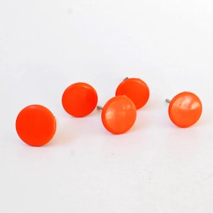 Könnte beinhalten: Fünf kleine, runde, leuchtend orangefarbene Schrankknöpfe mit glatter, glänzender Oberfläche. Jeder Knopf hat eine flache, kreisförmige Vorderseite und einen metallenen Schraubbolzen auf der Rückseite zur Befestigung an Möbeln oder Türen.
