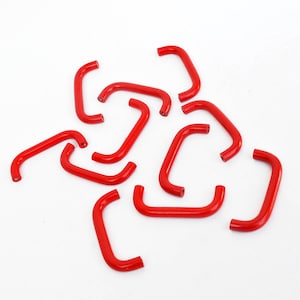 May include: A collection of bright red, curved metal staples scattered on a white background. Each staple has a rectangular shape with rounded corners, creating a U-shape. The staples are evenly spaced and appear to be new.