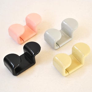 Può includere: Quattro ganci in plastica a forma di U in rosa pastello, grigio chiaro, nero lucido e crema. Ogni gancio presenta un design liscio e curvo con due estremità arrotondate e un ponte centrale, offrendo una forma funzionale per appendere.