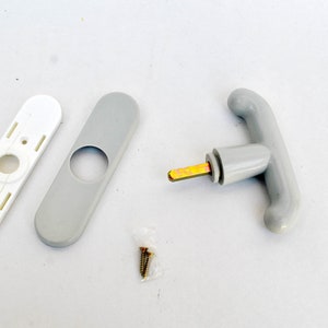Könnte beinhalten: Ein Satz an Beschlägen für einen Türgriff, bestehend aus einem grauen Griff, einer weißen und grauen Platte, einer Schraube und einer kleinen Tüte mit Schrauben.